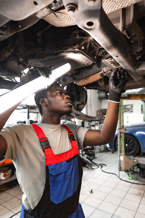 Young African mechanic in uniform working under the car in car service center close up